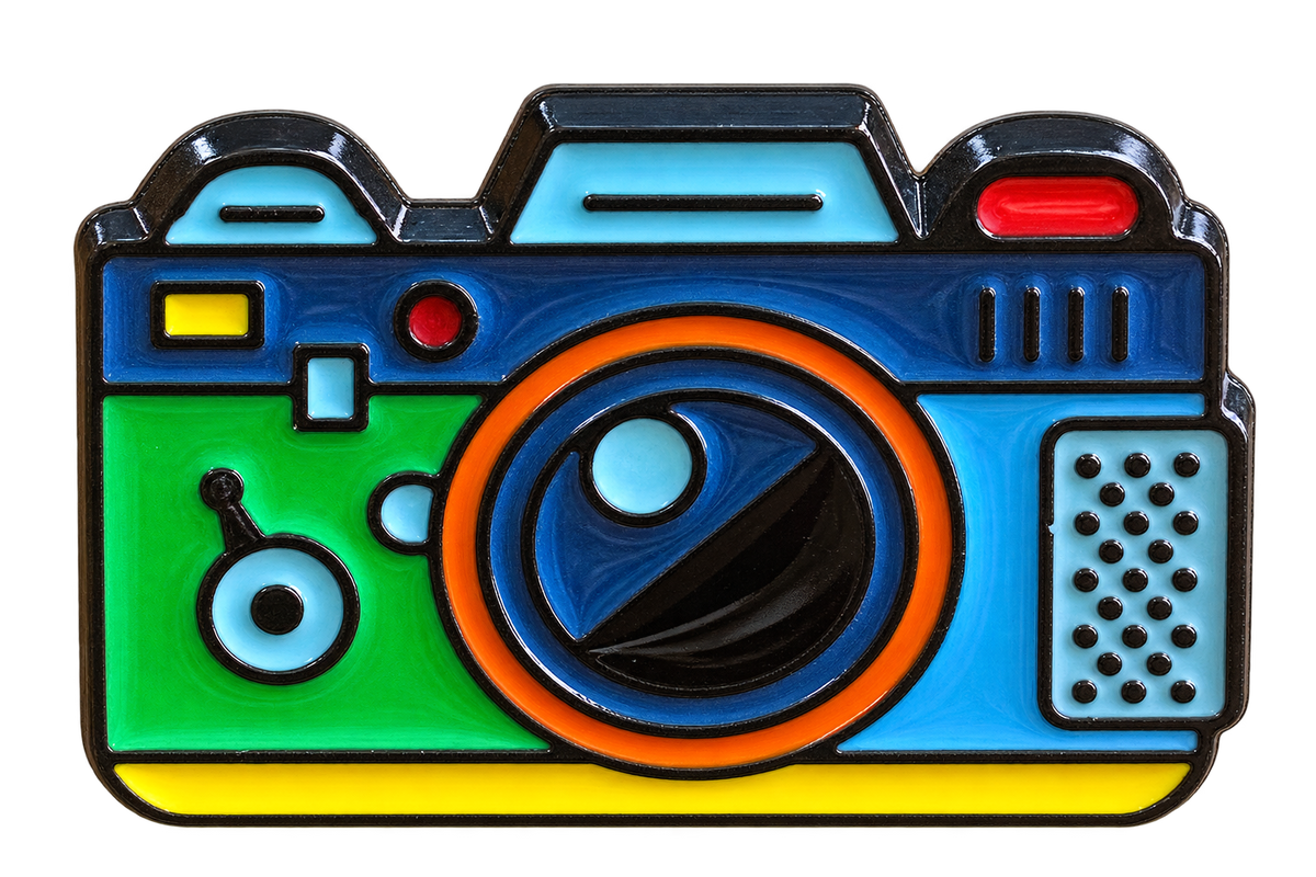 Kleurrijke camera emaille pin met blauwe en groene vlakken, oranje lens en gele accenten, voorzijde