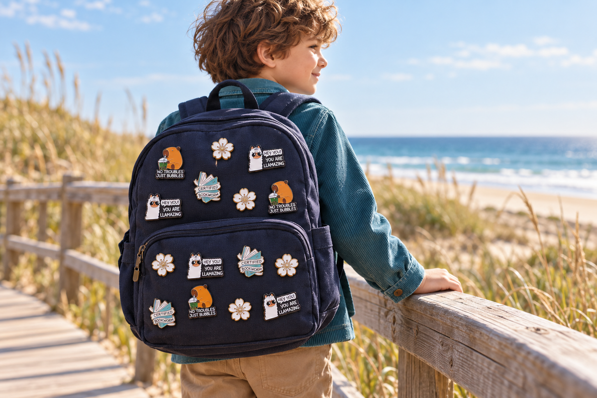 Anwendungsbeispiel: Kind am Meer mit dunkelblauem Rucksack und Capybara-, Lama-, Blumen- und Bücherwurm-Pins von Fasola